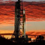 Silhueta do foguete SLS e da nave Orion, da missão Artemis II, na Plataforma de Lançamento 39B do Kennedy Space Center na Flórida. O céu está dramaticamente iluminado com nuvens em tons de vermelho e laranja pelo nascer do sol. Árvores e torres de transmissão aparecem em silhueta no primeiro plano