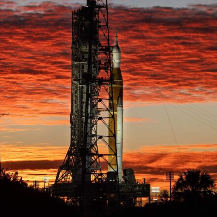 Silhueta do foguete SLS e da nave Orion, da missão Artemis II, na Plataforma de Lançamento 39B do Kennedy Space Center na Flórida. O céu está dramaticamente iluminado com nuvens em tons de vermelho e laranja pelo nascer do sol. Árvores e torres de transmissão aparecem em silhueta no primeiro plano